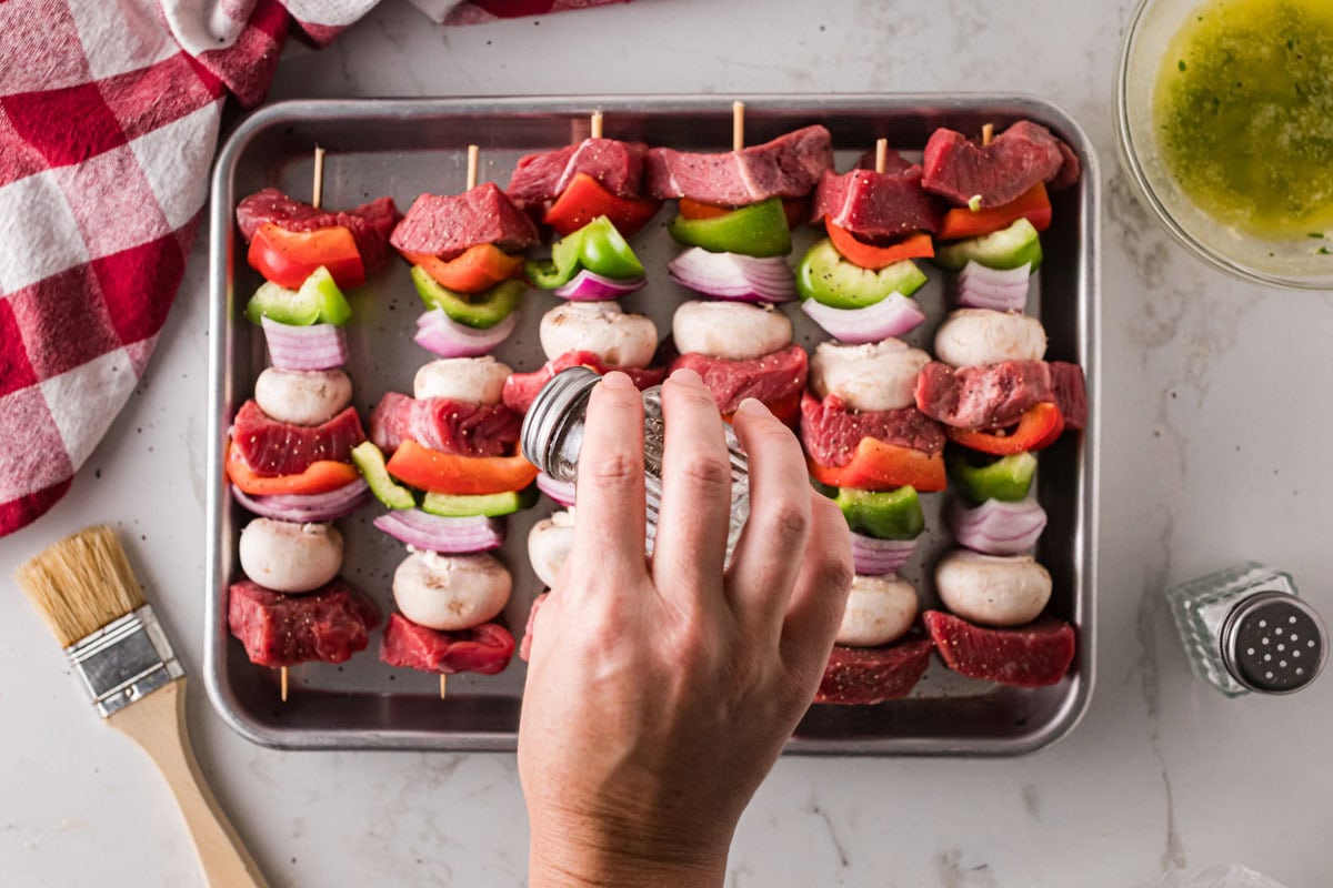 Sprinkling salt and pepper over steak kabobs.