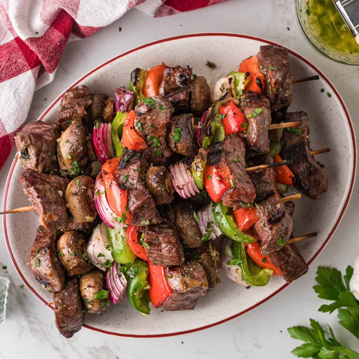 A white serving plate full a skewers with grilled steak kabobs with onions, peppers and mushrooms.