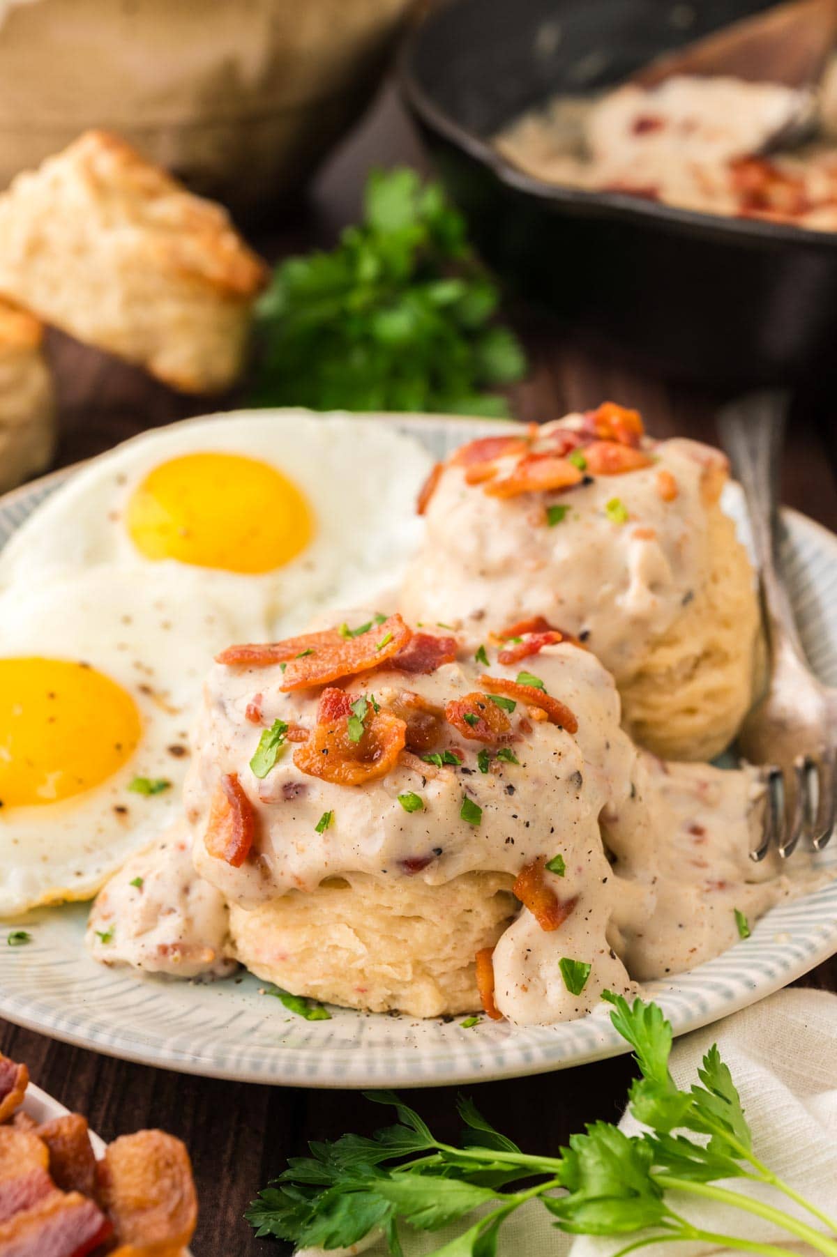 Bacon gravy on top of southern buttermilk biscuits topped with crumbled bacon, black pepper and a sprinkling of parsley on a plate with 2 fried eggs.