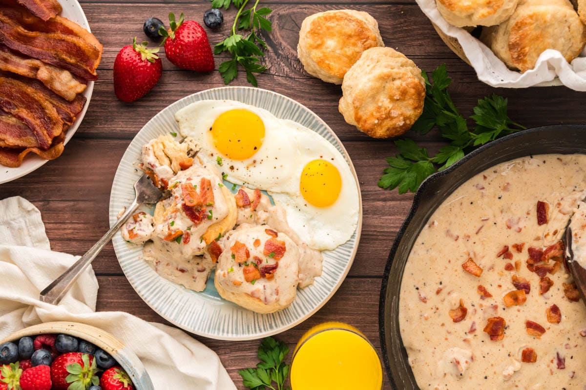 A breakfast tablescape with a plate of biscuits topped with bacon gravy and 2 eggs along with the cast iron skillet of bacon grease gravy with a plate of bacon and basket of biscuits on a wooden table.