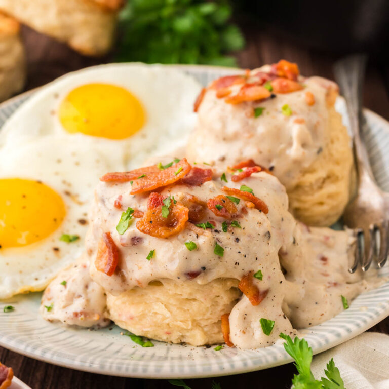 Bacon gravy on top of southern buttermilk biscuits topped with crumbled bacon, black pepper and a sprinkling of parsley on a plate with 2 fried eggs.