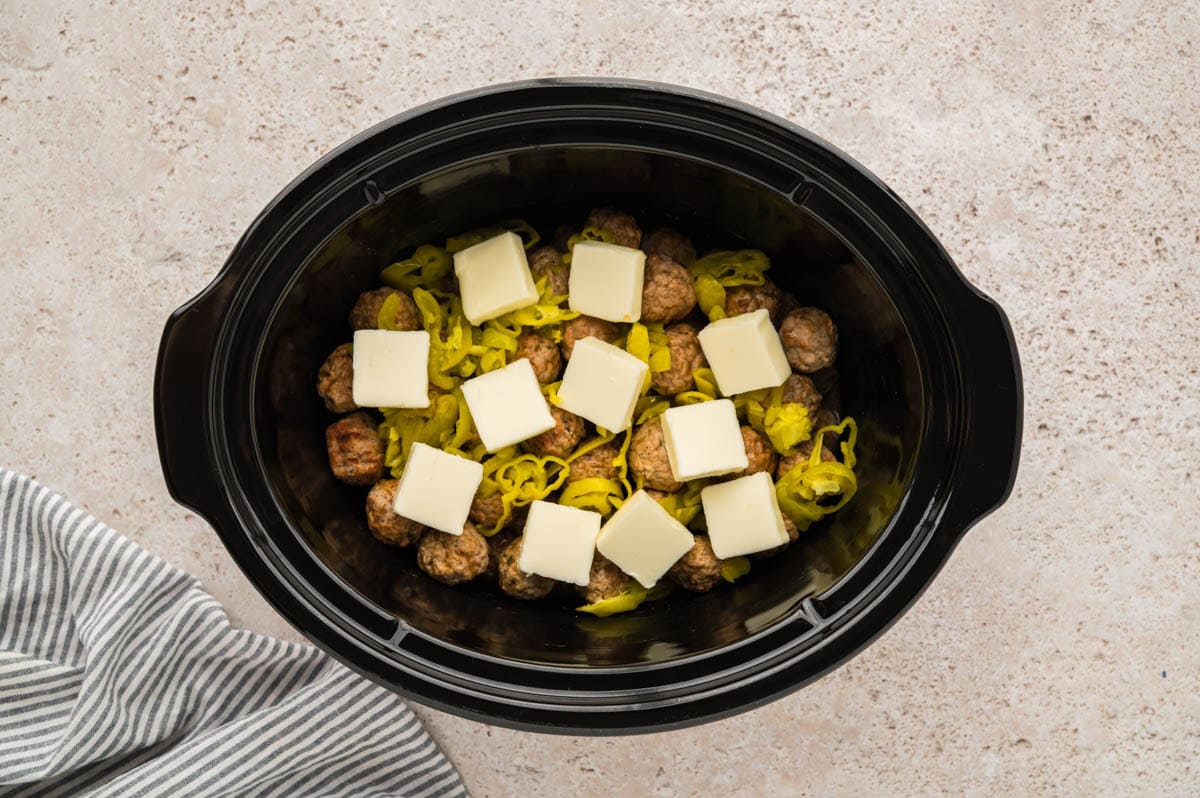Pats of butter placed on top of meatballs and pepperoncini peppers in a slow cooker.