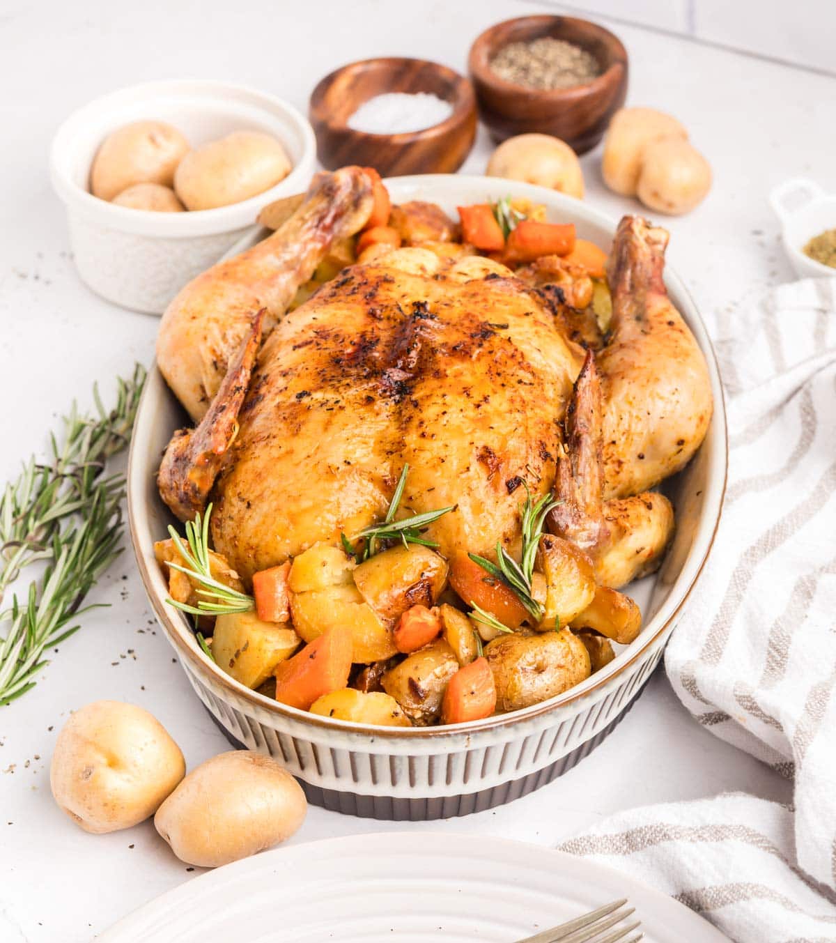 Roasted whole chicken in a serving dish with a side of carrots and potatoes.
