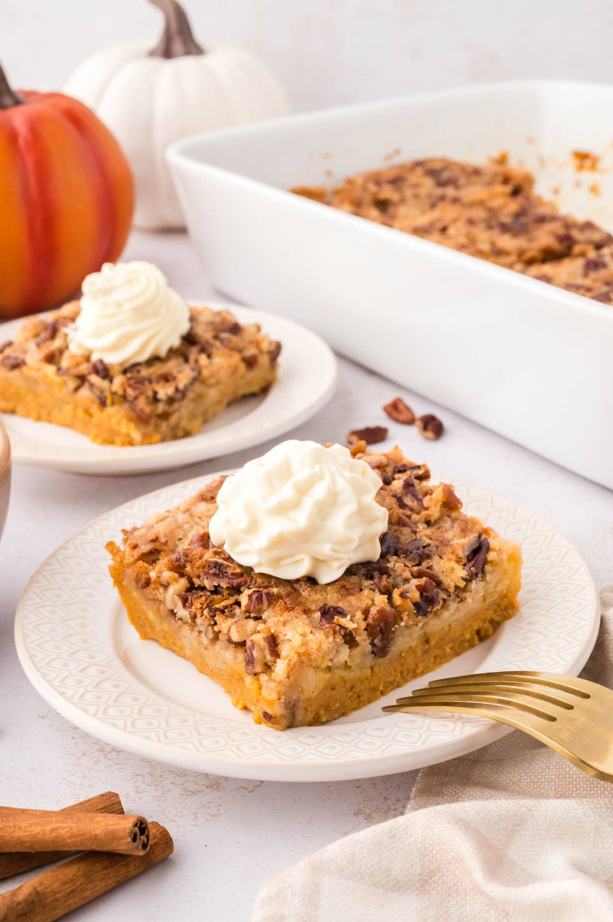Two white serving plates each with a slice of pumpkin dump cake topped with whipped cream and the whole casserole dish in the background.