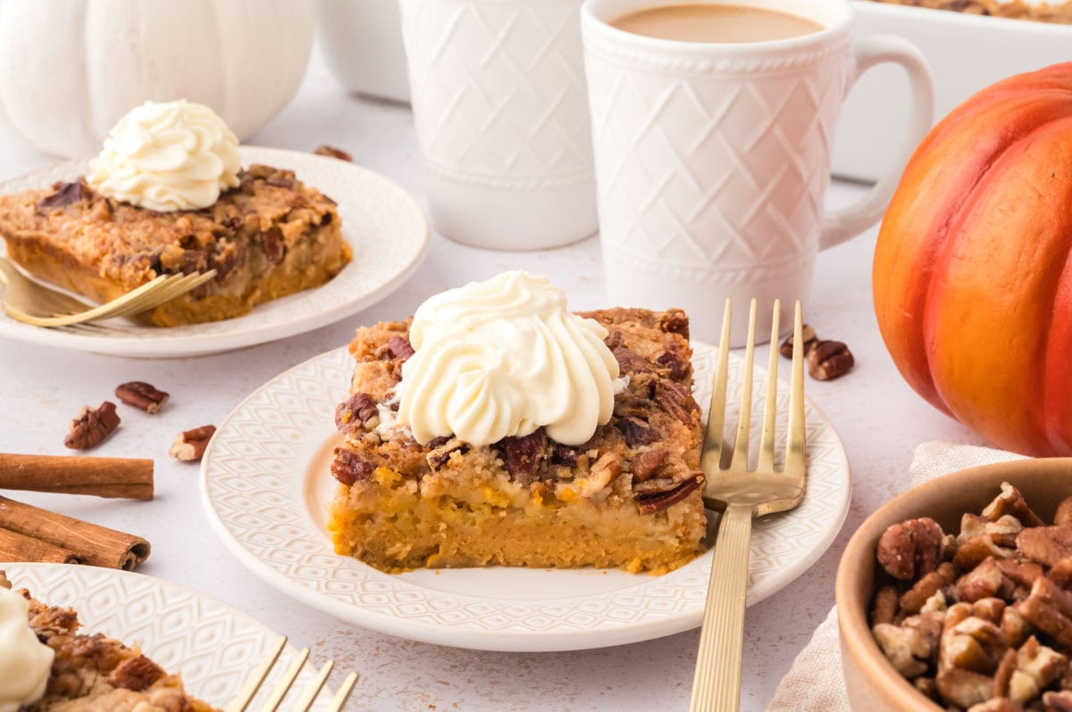Two white serving plates each with a slice of pumpkin dump cake topped with whipped cream and cups of coffee in the background.