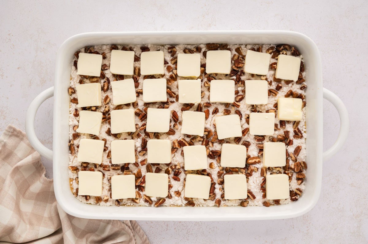 Sliced butter pats evenly distributed on top of the chopped pecan layer in the baking dish.