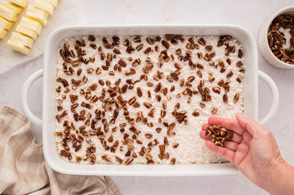 A hand sprinkling chopped pecan on top of the cake mix layer in the baking dish.