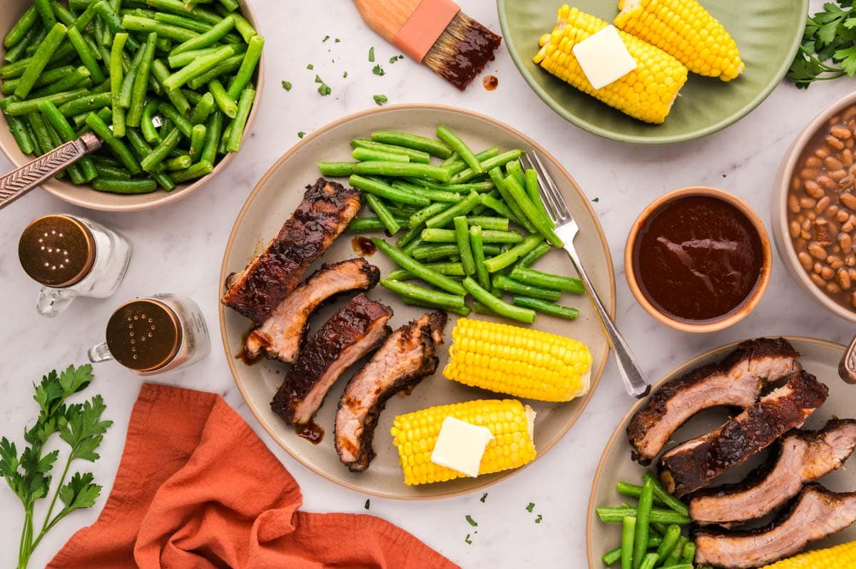 A tablescape with with a plate of sliced bbq baby back ribs, corn on cob, and green beans with serving bowls with baked beans, green beans and corn.