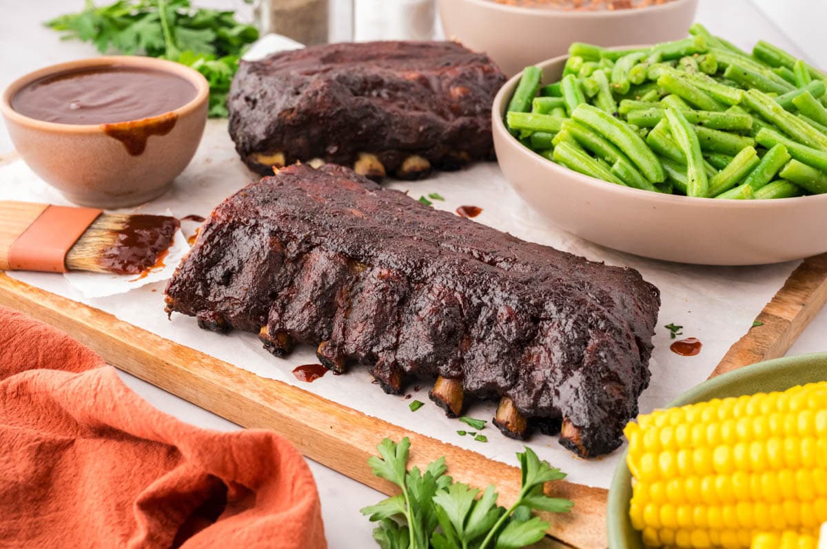 Two slabs of ribs on a wooden board with corn on the cob and a serving dish of bbq sauce.