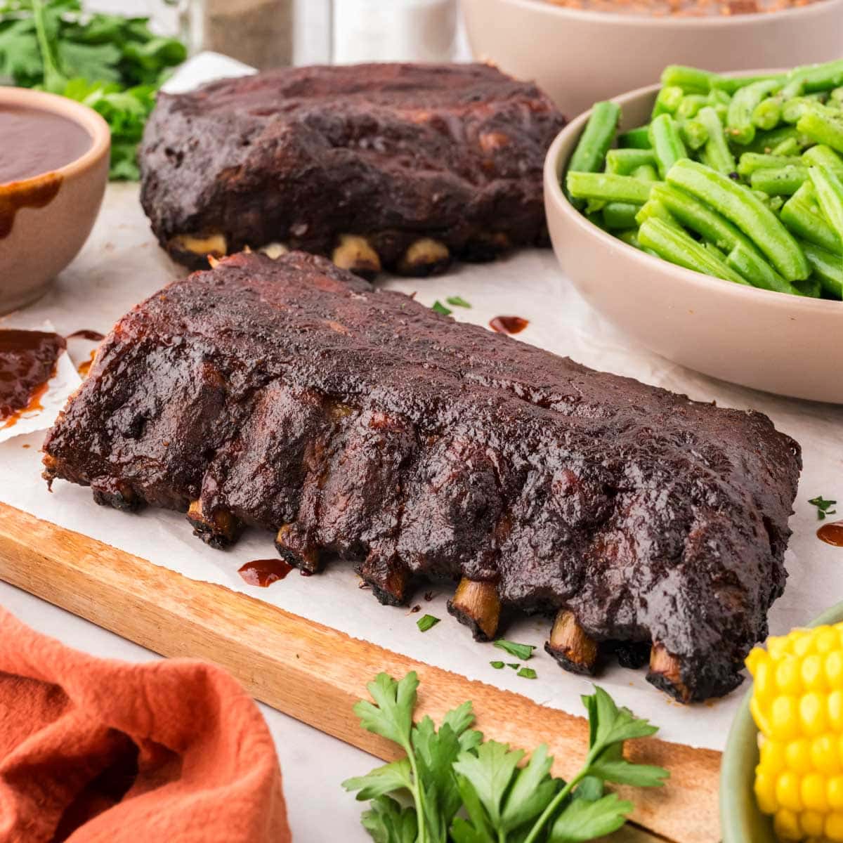 Two slabs of ribs on a wooden board with corn on the cob and a serving dish of bbq sauce.