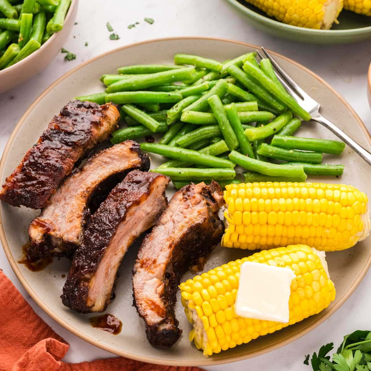 A plate of sliced smoked bbq baby back ribs, corn on cob, and green beans.