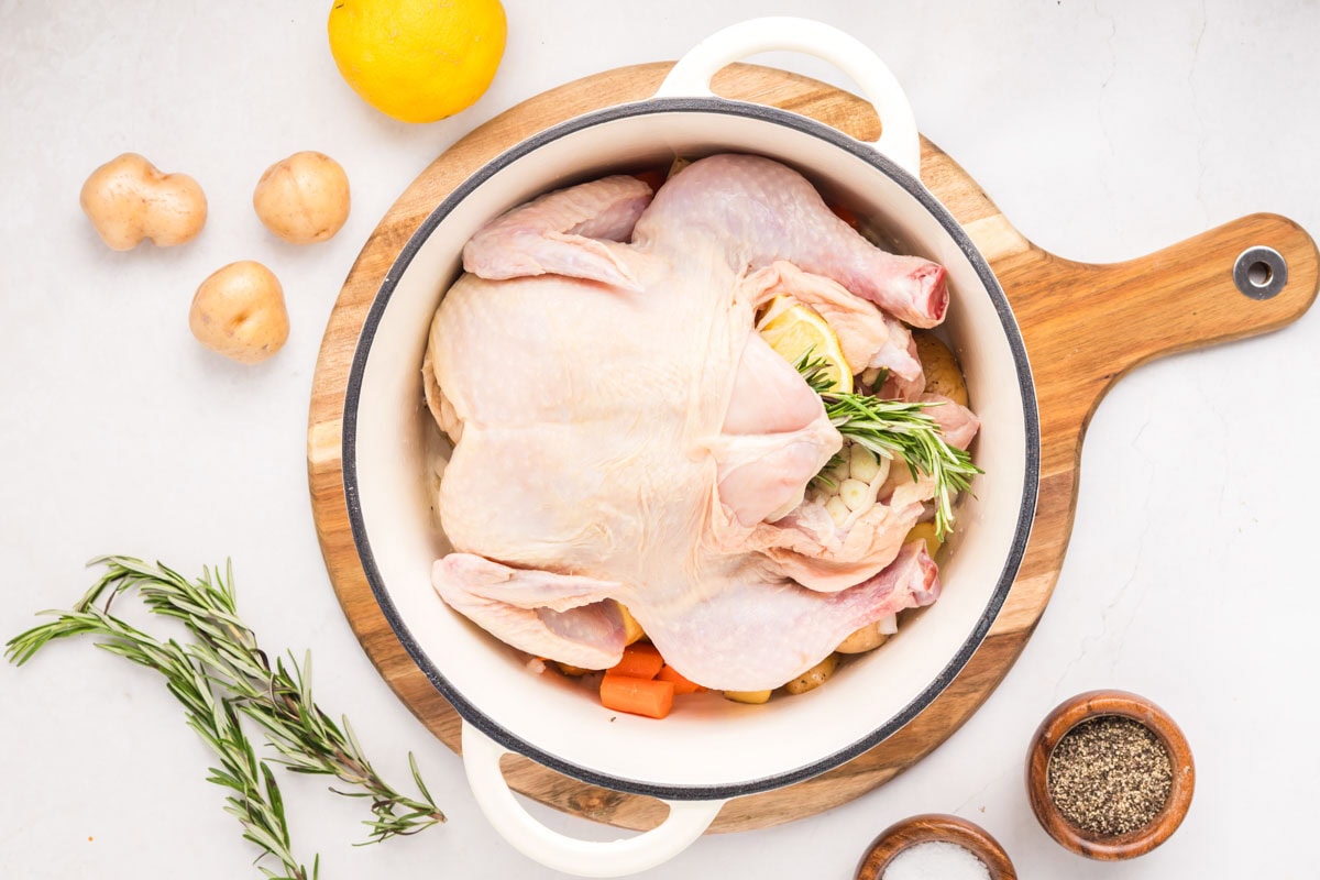 Whole raw chicken placed in the Dutch oven with potatoes and carrots.
